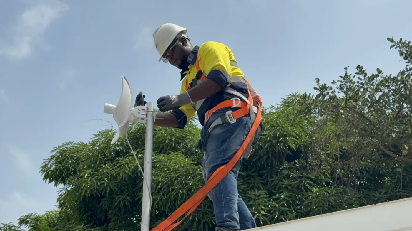 Kofi in a hard hat and harness installing a network dish on a rooftop